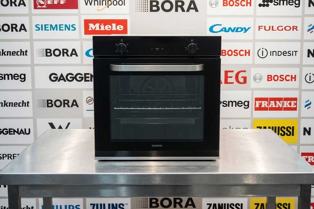 Ein Backofen steht auf einem Edelstahltisch vor einer Markenwand mit Logos bekannter Backofenhersteller.
