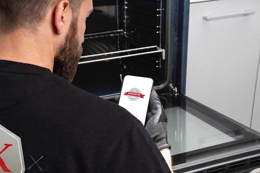 Blick über die Schulter eines Fachmanns, der während der Arbeit an einem Herd sein Smartphone nutzt. Auf dem Display ist das Logo für die Reparatur-Garantie zu sehen, was Sicherheit und Transparenz für den Kunden unterstreicht.