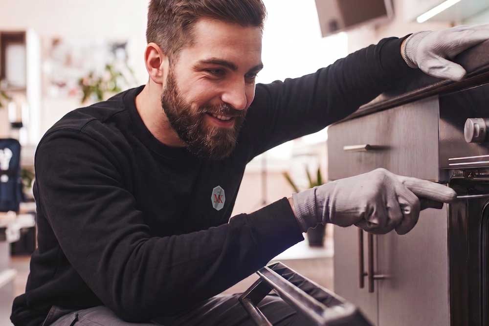 Ein freundlich lächelnder Techniker prüft die Mechanik eines Backofens in einer Kundenküche. Er trägt Schutzhandschuhe und zeigt auf ein Detail am Gerät, während er dem Kunden den Fortschritt der Reparatur erklärt.