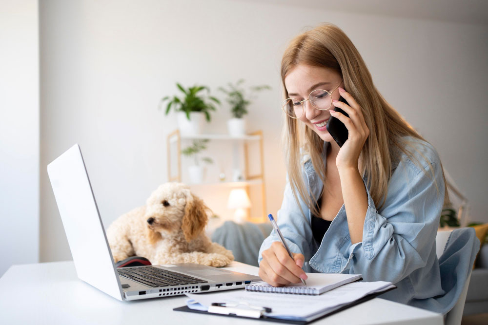 Frau sitzt an einem Schreibtisch, auf dem sich ein Laptop befindet, hält ein Blatt Papier in der Hand und telefoniert.
