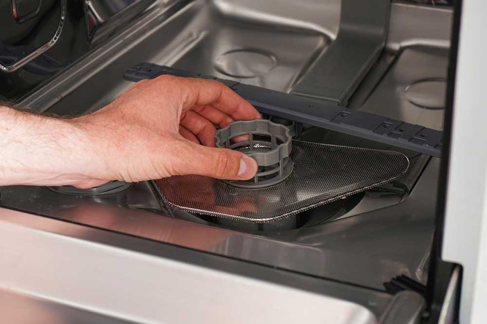 Nahaufnahme einer Hand, die das Filtersieb am Boden einer Geschirrspülmaschine zur Reinigung oder Wartung herausdreht. Das Bild verdeutlicht die Handhabung des mehrteiligen Filtersystems, welches für ein hygienisches Spülergebnis und die Langlebigkeit des Geräts regelmäßig gepflegt werden sollte.