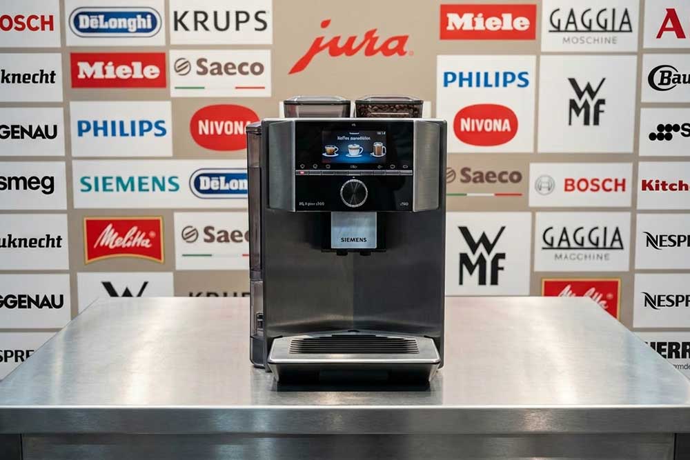 Ein moderner Kaffeevollautomat von Siemens steht auf einem Edelstahltisch vor einer Markenwand mit Logos bekannter Kaffeemaschinen-Hersteller wie Jura, DeLonghi, Miele, Philips und Saeco.