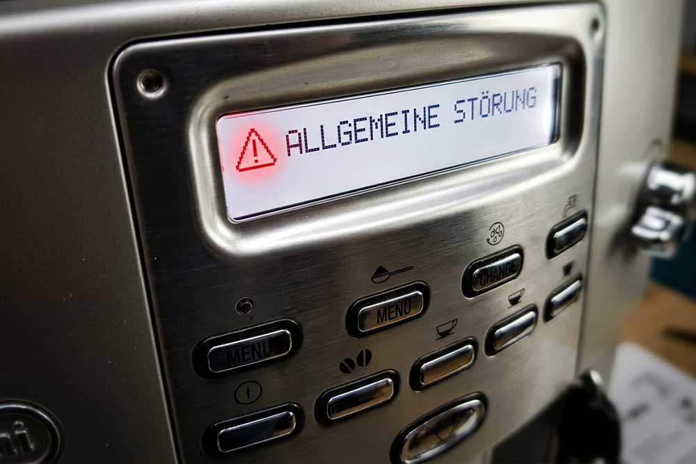 Nahaufnahme eines Kaffeemaschinen-Displays mit einem roten Warnsymbol und der Fehlermeldung 'ALLGEMEINE STÖRUNG' auf anthrazitfarbenem Gehäuse mit Bedientasten.