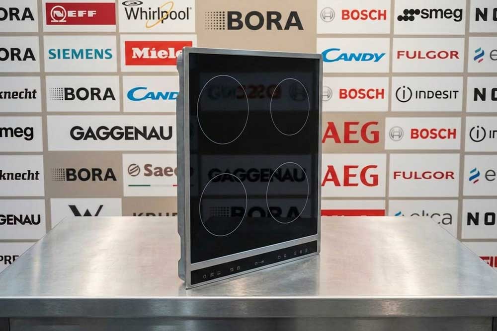 Ein modernes Kochfeld steht auf einem Edelstahltisch vor einer Markenwand mit Logos bekannter Kaffeemaschinen-Hersteller wie AEG, Bora, Indesit, Candy und Whirlpool.