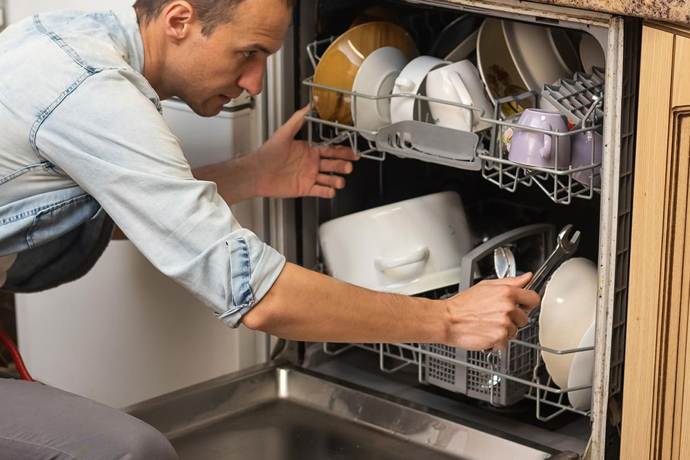 Techniker mit Schraubenschlüssel hockt vor einem geöffnetem Geschirrspüler