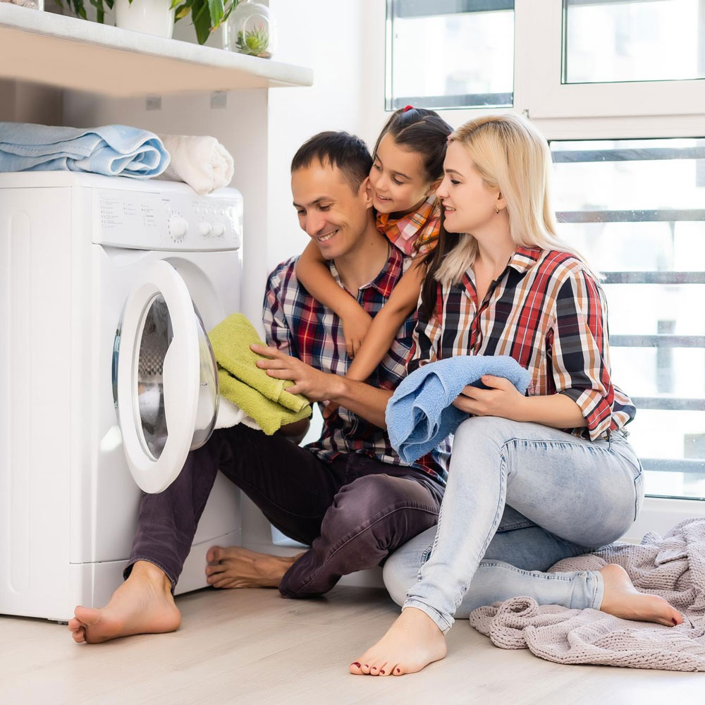 Mutter, Vater und Kind sitzen vor einer Waschmaschine und legen Handtücher zusammen.