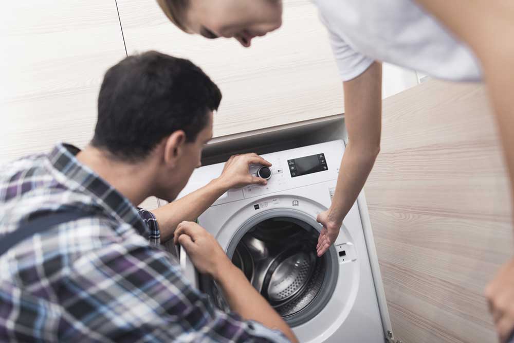 Ein Techniker erklärt einer Kundin die Fehlerdiagnose am Bedienfeld eines weißen Haushaltsgeräts in einer hellen Wohnung.